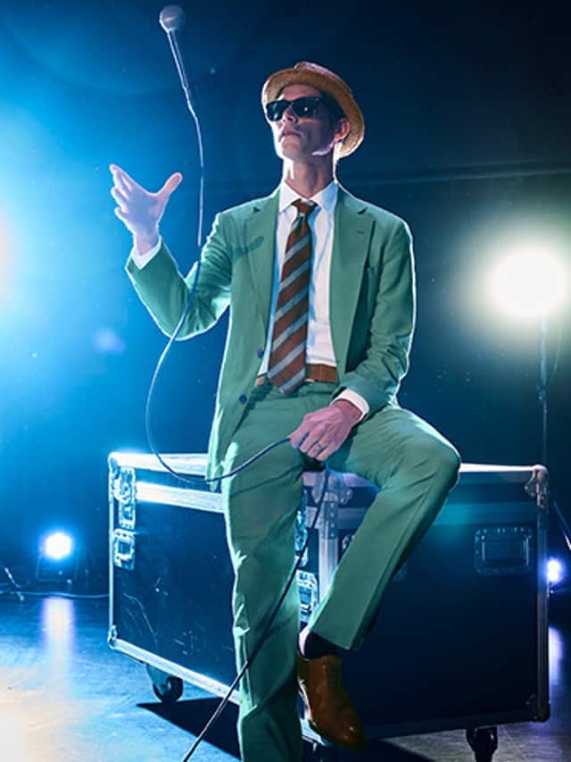 Auf dem Bild ist ein Mann zu sehen, der in einem mintgrünen Anzug mit gestreifter Krawatte, braunen Schuhen, Sonnenbrille und Strohhut auf einem Bühnenkoffer sitzt und ein Mikrofon hält.