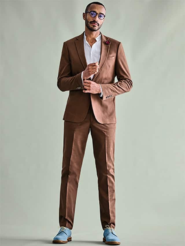 Ein Mann trägt einen braunen Anzug mit weißem Hemd und kombiniert ihn mit hellblauen Schuhen sowie einer Sonnenbrille.