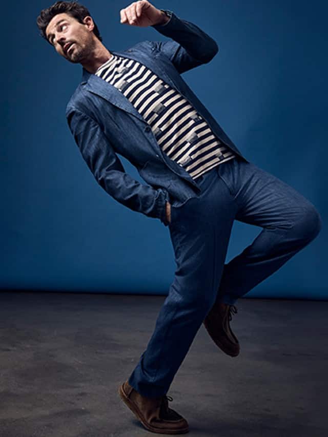 Ein Mann bekleidet mit einem Denim Anzug steht in einer akrobatischen Position vor einem blauen Hintergrund.