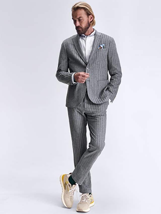 Ein Mann trägt einen grau gestreiften Anzug dazu helle Sneaker  und ein weißes Hemd darunter.