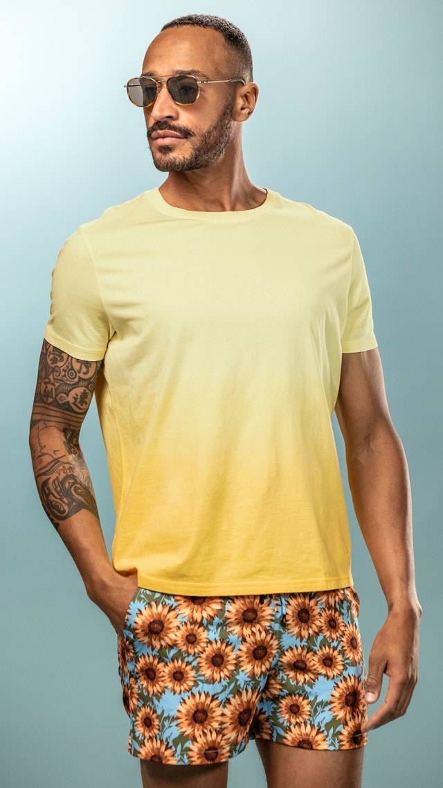 Ein Mann trägt ein gelbes T-Shirt und Shorts mit Sonnenblumenmuster, dazu eine Sonnenbrille.