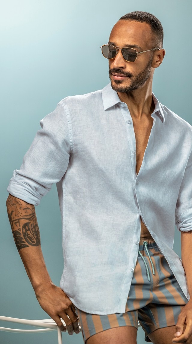 Ein Mann trägt ein hellblaues langärmliges Hemd, gestreifte Shorts in Blau und Braun sowie eine Sonnenbrille.