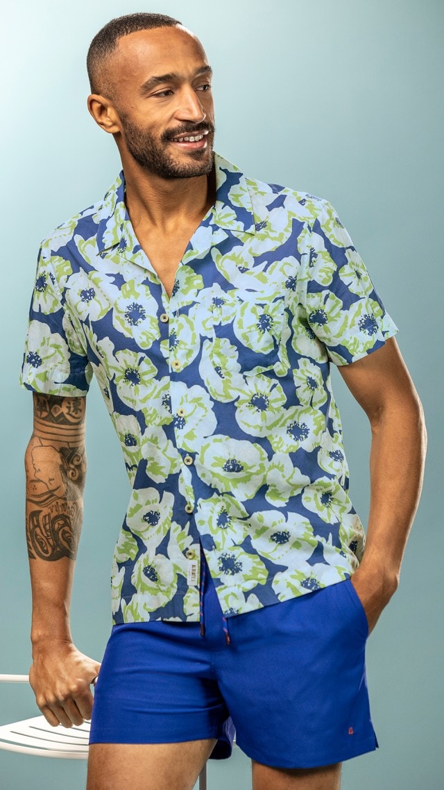 Ein Mann trägt ein kurzärmeliges Hemd mit blau-grünem Blumenmuster und blaue Shorts.