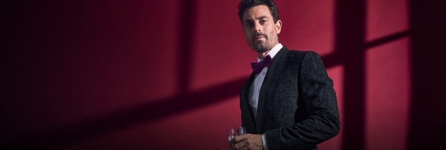 Ein Mann trägt einen eleganten, dunklen Anzug mit einem weißen Hemd und einer auffälligen magentafarbenen Fliege, während er ein Glas in der Hand hält. Ein Mann trägt einen eleganten, dunklen Anzug mit einem weißen Hemd und einer auffälligen magentafarbenen Fliege, während er ein Glas in der Hand hält.