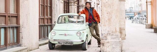 Ein Mann lehnt lässig an einem mintgrünen Oldtimer, während er in einer hellen Altstadtgasse steht.