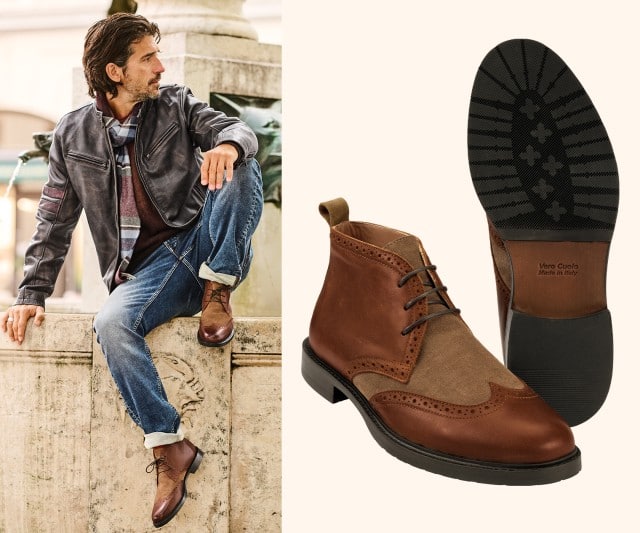 Auf dem Bild sitzt ein Mann lässig vor einem alten Industriegebäude, trägt eine braune Samtjacke mit Muster, eine Weste, ein Hemd, dunkle Jeans, braune Lederstiefel und eine Schirmmütze.