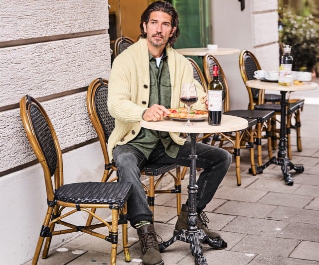 Ein Mann sitzt vor einem Straßencafé, trägt eine helle Strickjacke und isst Pizza bei einem Glas Rotwein.