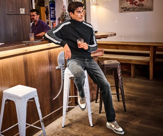 Ein Mann sitzt in einer Bar auf einem hohen Hocker, trägt einen dunklen Pulli mit Streifen an den Ärmeln und hält ein Getränk in der Hand.