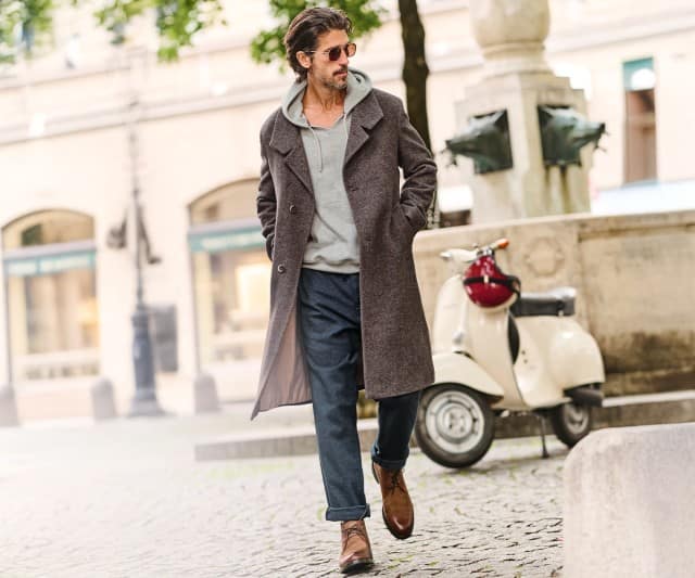 Ein Mann geht entspannt durch die Stadt und trägt dabei einen langen Mantel, einen Hoodie und braune Lederstiefel.