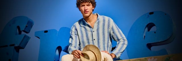 Eine Person in einem gestreiften Hemd und heller Hose hält einen Strohhut und posiert vor großen blauen Buchstabenformen im Freien bei sonnigem Wetter. Eine Person in einem gestreiften Hemd und heller Hose hält einen Strohhut und posiert vor großen blauen Buchstabenformen im Freien bei sonnigem Wetter.
