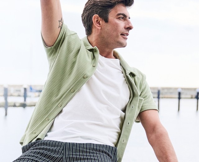 Auf dem Bild ist ein Mann zu sehen, der ein weißes T-Shirt, ein offenes hellgrünes, kurzärmliges Hemd und eine gestreifte Hose trägt.