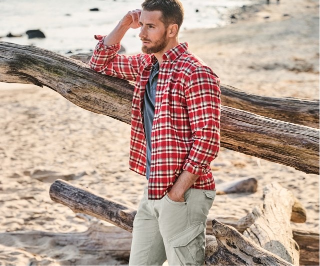 Auf dem Bild steht ein Mann am Strand, der ein rotes kariertes Hemd über einem dunkelgrauen T-Shirt sowie eine helle Cargohose trägt.