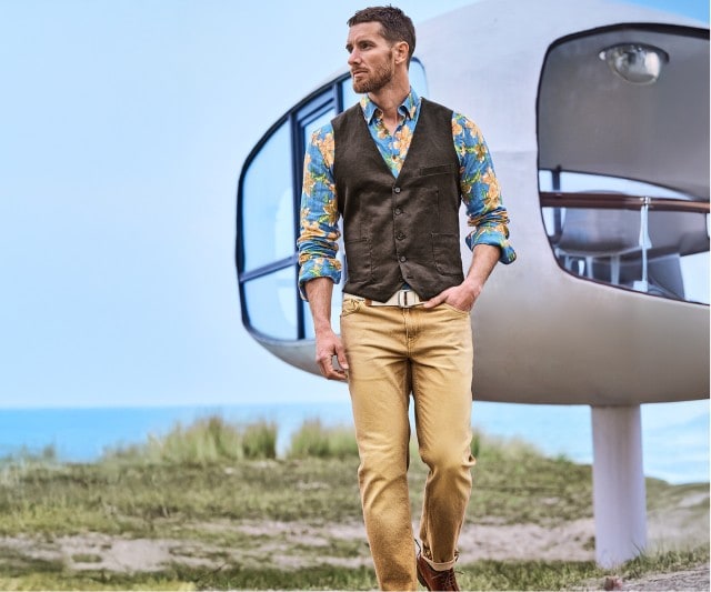 Auf dem Bild trägt ein Mann ein blaues Hemd mit buntem Blumenmuster, eine dunkle Weste sowie eine beige Hose mit hellem Gürtel.