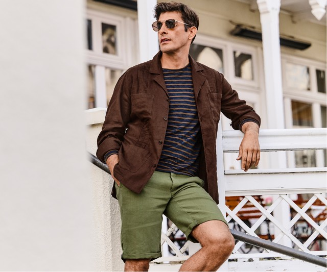 Auf dem Bild trägt ein Mann eine braune Hemdjacke über einem dunkelblau-oliv gestreiften T-Shirt sowie olivgrüne Shorts und eine Sonnenbrille.