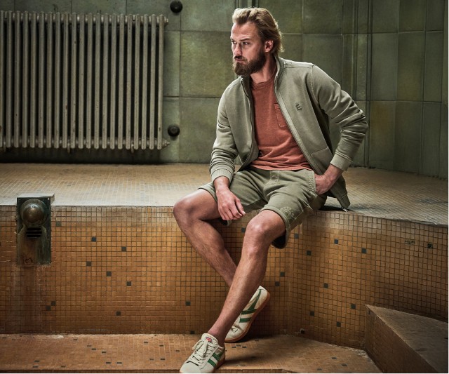 Auf dem Bild sitzt ein Mann in einem leeren Becken und trägt eine khakifarbene Trainingsjacke, ein rostfarbenes T-Shirt, dazu passende khakifarbene Shorts und weiße Sneaker mit grünen Streifen.