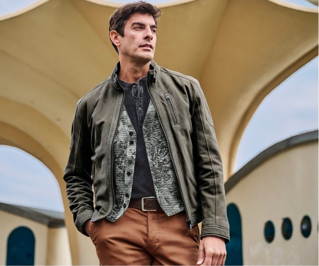 Auf dem Bild trägt ein Mann eine olivgrüne Jacke über einer grau gemusterten Weste und einem dunklen Henley-Shirt sowie eine braune Hose.