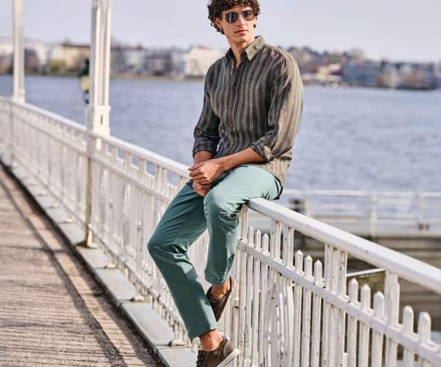Eine Person mit Sonnenbrille sitzt auf einem weißen Geländer an einer Uferpromenade und trägt ein gestreiftes Langarmhemd, eine hellgrüne Hose sowie dunkle Freizeitschuhe vor einer Wasser- und Stadtkulisse.