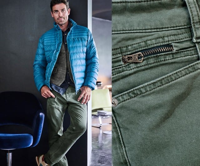 Ein Mann steht lässig an einer Wand, trägt eine blau gesteppte Jacke und olivgrüne Hose, die rechts im Detail mit einem Nahzoom des Reißverschlusses gezeigt wird.