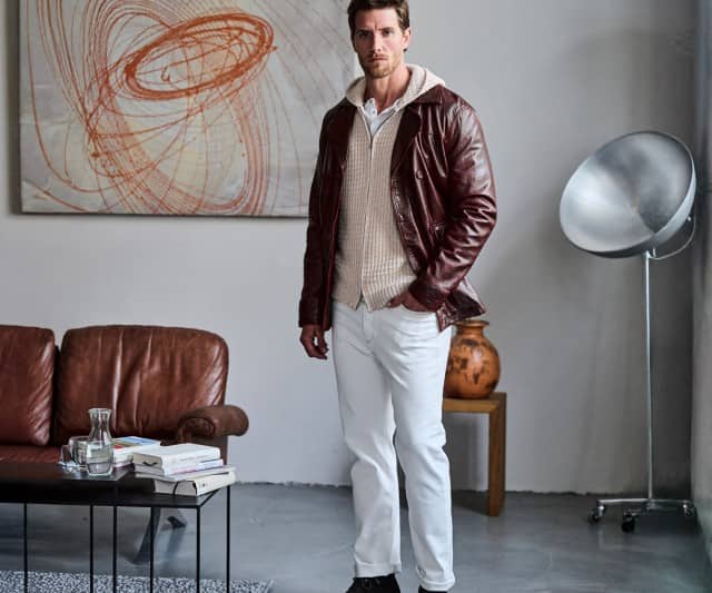 Ein Mann steht in einem modern eingerichteten Wohnzimmer und trägt eine dunkelrote Lederjacke über einem beigen Hoodie mit weißen Jeans.