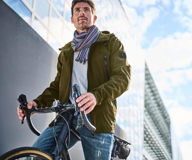 Ein Mann steht mit einem Fahrrad vor einem modernen Gebäude und trägt eine olivgrüne Jacke, einen hellen Pullover, Jeans und einen bunt gestreiften Schal.