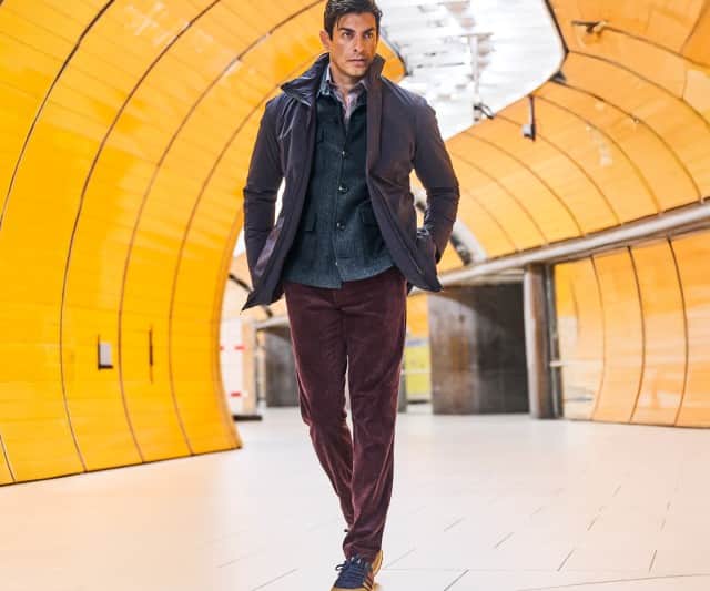 Ein Mann geht durch einen leuchtend gelben U‑Bahn‑Tunnel und trägt eine dunkle Jacke, einen grauen Blazer und bordeauxrote Hose.