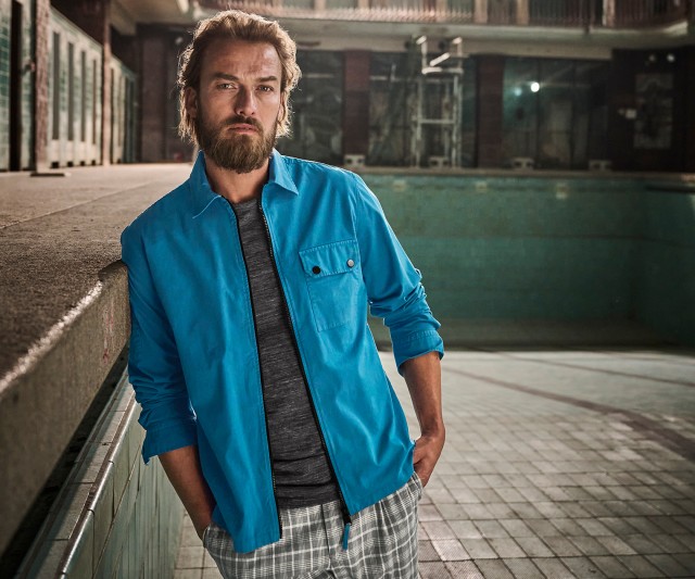 Auf dem Bild steht ein bärtiger Mann in lässiger Kleidung (hellblaue Jacke, graues T-Shirt und karierte Hose) in einem leeren, verlassenen Schwimmbecken und lehnt mit einer Hand an der Kante.