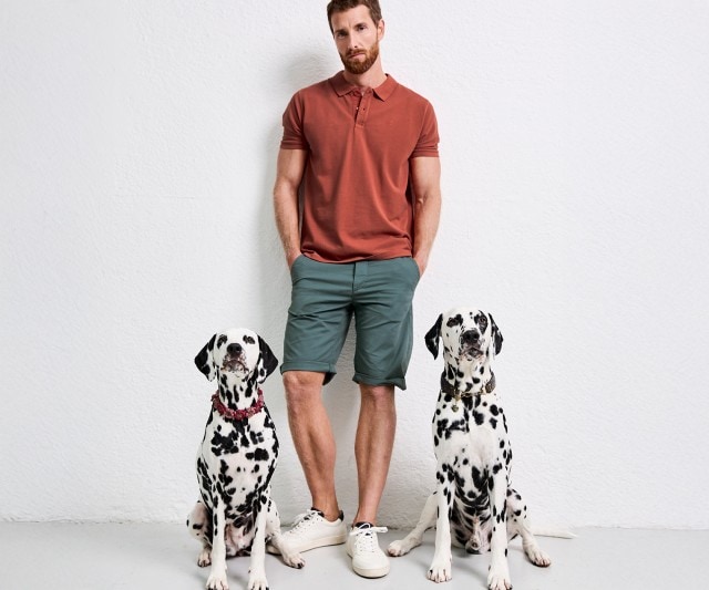 Auf dem Bild steht ein Mann mit rötlich-braunem Poloshirt, grünen Shorts und weißen Sneakern zwischen zwei Dalmatinern, die auf dem Boden sitzen und in die Kamera blicken.