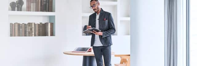 Ein Mann trägt einen strukturierten, dunkelgrauen Anzug mit hellgrauem Shirt, Einstecktuch und Brille, während er in einem Notizbuch schreibt.