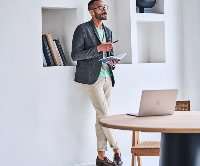 Ein Mann trägt einen dunkelgrauen Blazer über einem mintgrünen T‑Shirt und einer hellen Weste, kombiniert mit beigefarbenen Hosen und braunen Loafern.