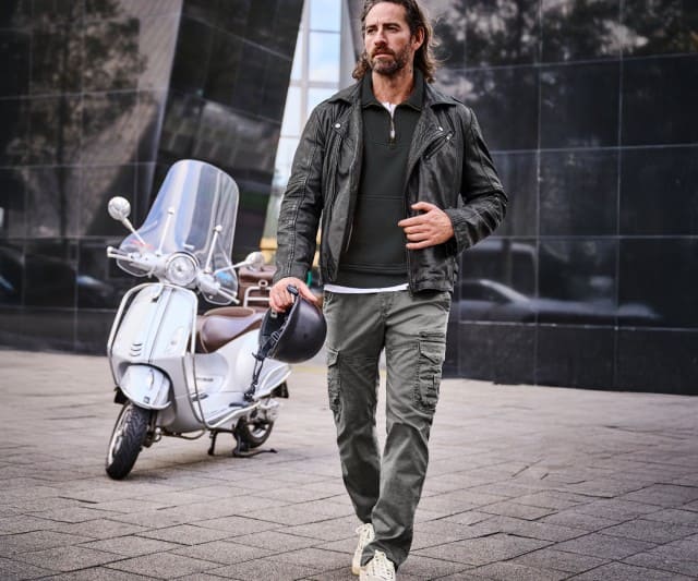 Ein Mann trägt eine schwarze Lederjacke über einem dunkelgrünen Zip‑Sweater, kombiniert mit grauen Cargo‑Hosen und hellen Sneakern.