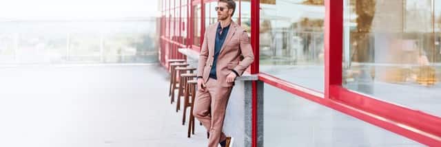 Ein Mann trägt einen hellbraunen Anzug mit passender Hose und Blazer, kombiniert mit einem dunkelblauen, gemusterten Hemd und Sonnenbrille.