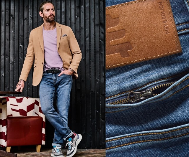 Ein Mann trägt ein beigefarbenes Sakko über einem hellvioletten T‑Shirt, kombiniert mit einer blauen Jeans und sportlichen Sneakers.