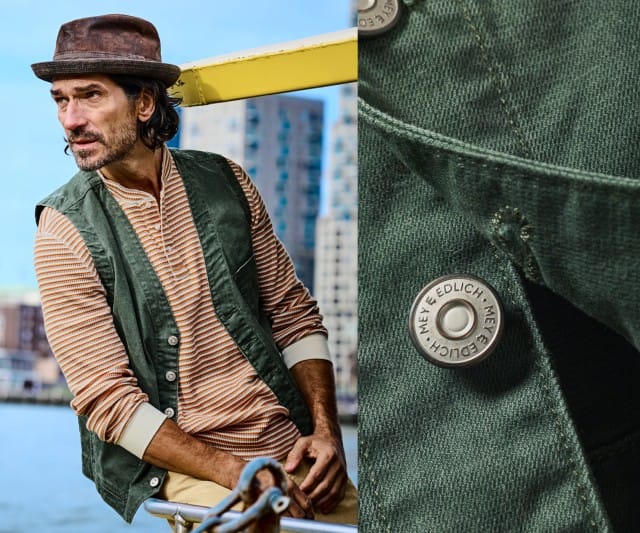 Ein Mann trägt ein grün gefärbtes, ärmelloses Weste‑Gilet über einem langärmeligen, orange‑cremefarben gestreiften Henley‑Shirt und kombiniert es mit einer beigefarbenen Hose.