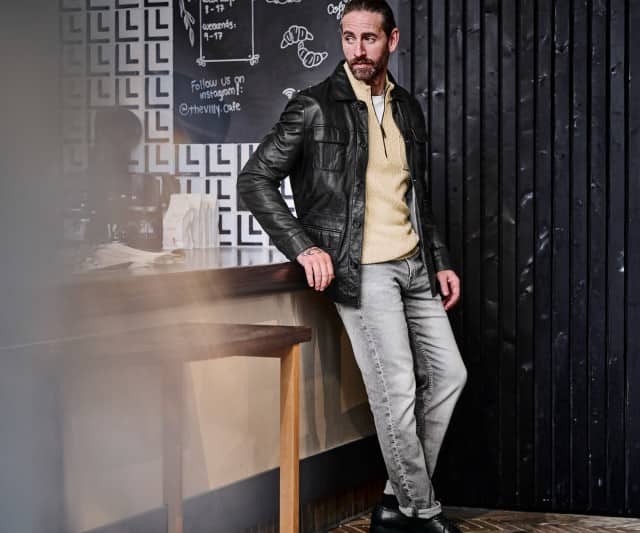 Ein Mann trägt eine schwarze Lederjacke über einem beigefarbenen Strickpullover mit kurzem Zipper, kombiniert mit hellgrauen Jeans und schwarzen Schuhen.