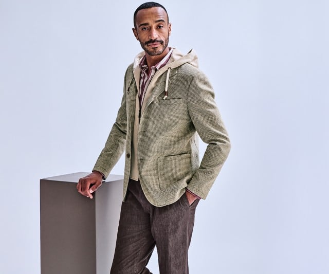 Ein Mann trägt ein hellgrünes, fein gemustertes Sakko mit eingenähter Kapuze über einem gestreiften Hemd und kombiniert es mit einer dunkelbraunen Hose.