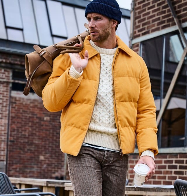 Ein Mann trägt eine gelbe Steppjacke über einem cremefarbenen Strickpullover, eine braune Hose, eine dunkelblaue Mütze und hat eine braune Tasche über der Schulter sowie einen Kaffeebecher in der Hand. Ein Mann trägt eine gelbe Steppjacke über einem cremefarbenen Strickpullover, eine braune Hose, eine dunkelblaue Mütze und hat eine braune Tasche über der Schulter sowie einen Kaffeebecher in der Hand.