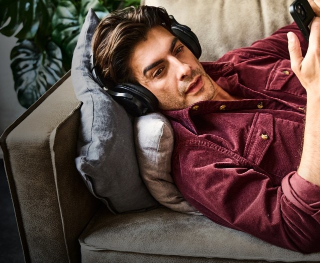 Ein Mann liegt entspannt auf einem Sofa und trägt ein weinrotes Cordhemd mit hochgekrempelten Ärmeln sowie eine beige Hose.