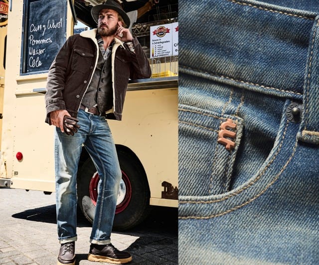 Ein Mann trägt eine braune Cordjacke mit heller Lammfellfütterung über einem Hemd und einer Weste, dazu eine hellblaue Jeans mit Aufschlag, braune Lederschuhe, Handschuhe und einen Hut.