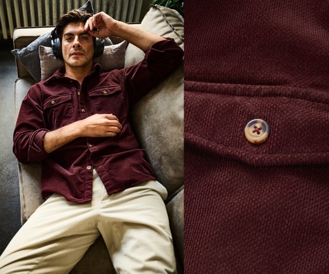 Ein Mann liegt entspannt auf einem Sofa und trägt ein weinrotes Cordhemd mit Knopfleiste und Brusttaschen sowie eine helle Hose.