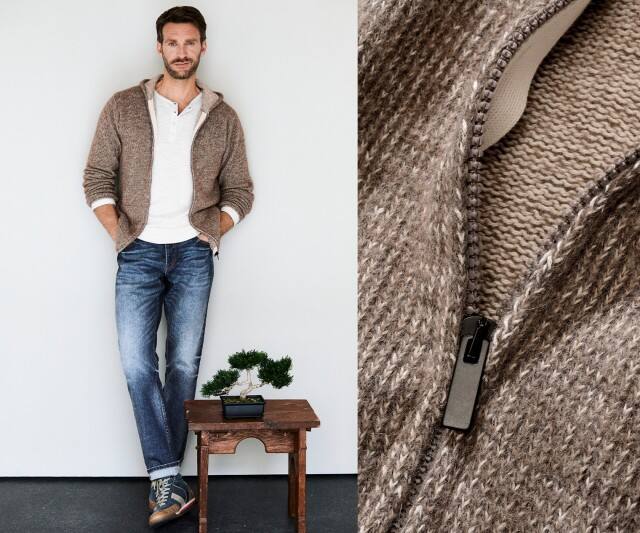 Ein Mann trägt eine beigefarbene Strickjacke mit Kapuze über einem weißen Henley-Shirt, dazu eine blaue Jeans und sportliche Sneaker.