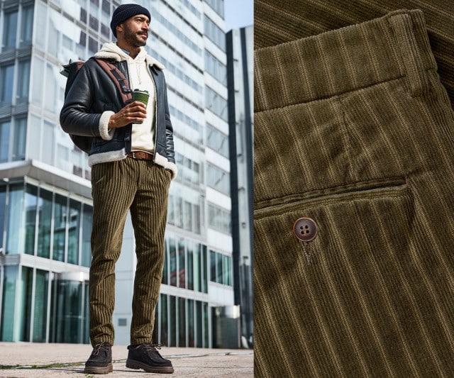 Ein Mann trägt eine dunkelblaue Fliegerjacke mit heller Lammfellfütterung über einem cremeweißen Hoodie, dazu eine olivgrüne Cordhose, dunkle Schuhe, eine Strickmütze und einen Rucksack.