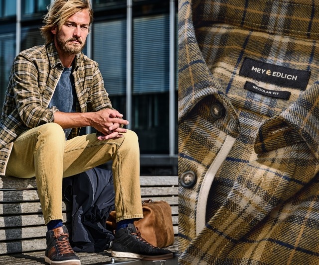 Ein Mann trägt ein gelb-braun kariertes Hemd über einem dunkelblauen T-Shirt, kombiniert mit einer hellgelben Hose und schwarzen Sneakers mit braunen Details.