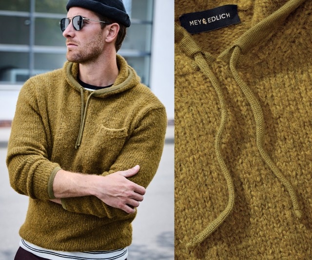 Ein Mann trägt einen senfgelben Strickpullover mit Kapuze und Kordelzug, kombiniert mit einem schwarzen T-Shirt darunter sowie einer schwarzen Mütze und Sonnenbrille.