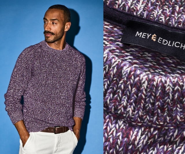 Ein Mann trägt einen grob gestrickten Pullover in einer lilafarbenen Melange-Optik, kombiniert mit einer weißen Hose und einem braunen Ledergürtel.