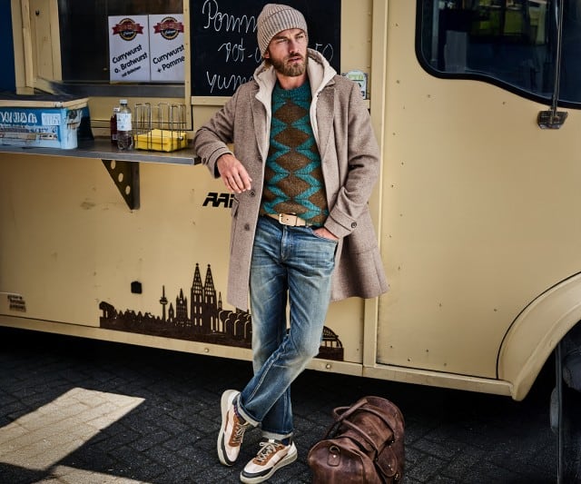 Ein Mann trägt einen beigen Mantel über einem grünen Pullover mit braunem Zickzackmuster, eine hellblaue Jeans mit Aufschlag, beigefarbene Sneaker und eine Strickmütze.