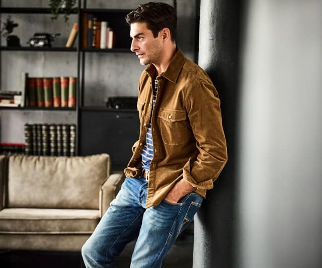 Ein Mann trägt ein braunes Cordhemd mit Brusttaschen über einem blau-gelb gestreiften Shirt und einer blauen Jeans.