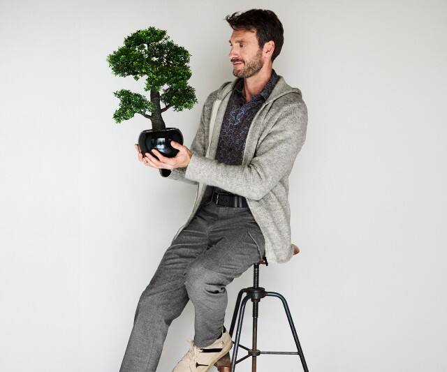 Ein Mann trägt eine hellgraue Strickjacke mit Kapuze über einem gemusterten Hemd, dazu eine graue Stoffhose und beigefarbene Schuhe.