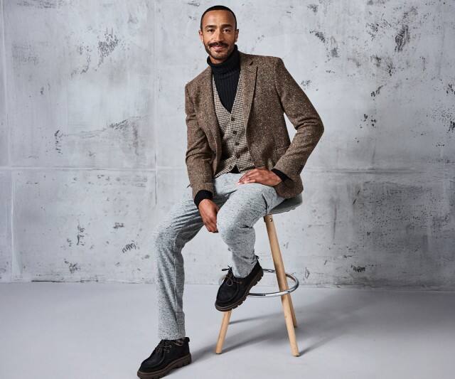 Ein Mann trägt ein braunes Sakko über einer gemusterten Weste und einem schwarzen Rollkragenpullover, dazu eine hellgraue Hose und dunkle Schuhe.