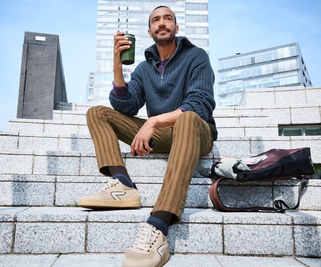 Ein Mann trägt einen dunkelblauen Strickpullover mit halbem Reißverschluss über einem violetten Shirt, dazu eine braun gestreifte Hose und beigefarbene Sneaker.
