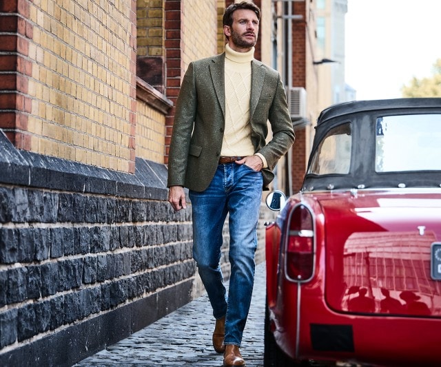 Ein Mann trägt einen olivgrünen Sakko über einem gelben Rollkragenpullover mit Strickmuster, kombiniert mit einer blauen Jeans und braunen Lederstiefeln.